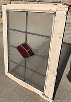 Front view showing the window with clear textured glass panes and contrasting red diamond accent, wood frame with chipped white paint.