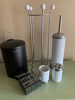 Full view of bathroom lot showing black mini waste bin, toilet brush and holder, cup, toothbrush holder, toilet paper holder, and mirrored box.