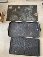 Photo of 2 black plastic boot trays and 1 black rubber snowman door mat placed on concrete floor