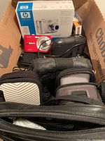 Box containing multiple cameras, cases, and cables: boxed HP Photosmart M627, red Kodak camera, black Kodak Cameo, and several black and gray cases