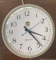 Front view of round electric wall clock with white face, black numerals and hands, blue second hand, metal rim.