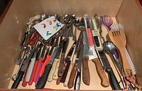 Top view of drawer containing assorted kitchen knives with various colored plastic and wooden handles, salad servers, and metal utensils