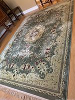 Full view of the large area rug laid out on hardwood floor showing its floral pattern and beige border with fringe edges.