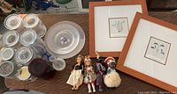 Shows assortment of glass jars, plates, framed botanical prints with wooden frames, and four dolls arranged on a surface.
