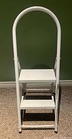 Front view of the white metal 2-step stool showing the handle and step platforms