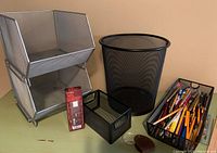 Photo showing two silver metal mesh stacking storage units, black metal mesh waste basket, black mesh drawer organizer, magnifying glass, and a transparent divided container holding pens, pencils, and small tools.