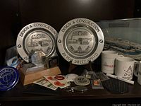 Photo showing two large Design & Construction 20 Year Safety Award plates with engraved plant images, assorted memorabilia including ceramic mugs, Zippo lighter, pins, patches, keychain, pen, buttons, paperweight, and stickers.