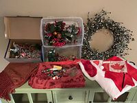 Photo showing various holiday decorative items arranged on top of a green sideboard, including wrapped red tablecloth, tasselled gold and crimson runner, two plastic bins containing greenery and red berries, a door wreath with frosted branches, ornament cheese spreaders, and a Canada flag towel folded on the right side.