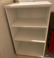 Front view of white shelving unit, showing three shelves and peg holes for adjustable shelves.