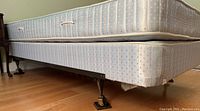 Side angled view showing metal bed frame on wheels below box spring and mattress, highlighting the frame structure and wheels.