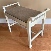 Side angled view showing off-white painted wooden stool with light brown fabric cushion, decorative handles/legs, and floor placement.