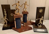 Photo showing four vintage bowling trophies with gold-tone figurines and dark wood bases, displayed together on a floor against a wood-paneled wall. Three female figures, one male, with plaques removed.