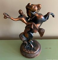 Front angle showing cowboy with hat and horse in rearing pose, chain on bridle, detailed saddle, and round base with decorative border and felt bottom