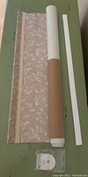 Rolled vinyl window blind with beige and white leaf/floral pattern, mounting bracket, and screws on green surface