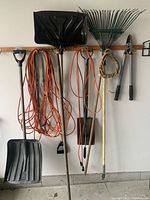 Full view of hanging yard and garden tools including rakes, shovels, spade, clippers, and extension cords hung on wall