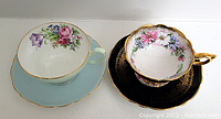 Two cups with their matching saucers, displayed side by side on a white surface, showing floral patterns and gold trim. One set is blue floral by Royal Stafford, the other black floral with gold details by Foley.