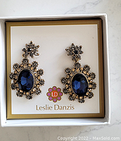 Pair of Leslie Danzis earrings with large oval blue faceted stones surrounded by small bead-like accents in a gold-tone metal, displayed on Leslie Danzis card inside a box.