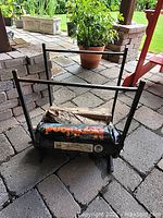 Front angled view of black metal fireplace log rack with firewood, showing wear and rust on frame and plastic wrapped wood