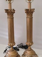 Pair of gold-colored metal column lamps with fluted shafts and Corinthian capitals, showing full height and cords.