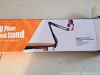 Box front with image of the phone desk stand mounted on a desk holding a smartphone, showing arm structure and clamp.
