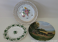 Three decorative plates displayed together on a white surface showing distinct styles: pierced floral Limoges plate, ivy-leaf Royal Albert plate, and painted Dresden plate with a hunting scene.