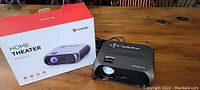Close view of Bomaker Home Theater projector next to its original box, showing compact design and branding on top.
