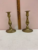 Pair of brass candle holders placed in front of a pink wall with a ruler behind them showing height approximately 7 inches.