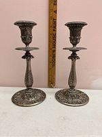 Pair of two silver plated candle holders with intricate floral and scrollwork design, shown side by side against a wall with a measuring stick behind them to show height.