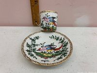 Cup and saucer with colorful bird painting and gold trim placed on white surface