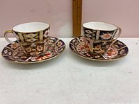 Photo shows two Royal Crown Derby cups each paired with a matching saucer. The cups and saucers have detailed Imari style patterns in cobalt blue, red, white and gold.