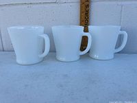 Three white opaque milk glass Fire King coffee mugs placed against white wall, all with handles visible. Wooden ruler in background for size context.