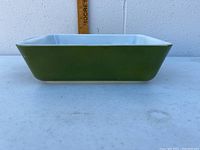 Side view of a green rectangular Pyrex casserole dish with light blue interior, placed on a white surface against a gray wall showing measurement ruler for scale.