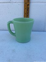 Front angle photo of single jadeite green glass Fire King mug with handle, measuring approximately 4 inches tall against a wooden ruler.