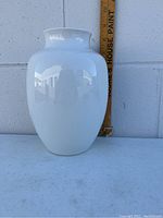 Front view of tall, glossy white porcelain vase with smooth finish and flared neck, against a wall with measuring stick behind for scale.