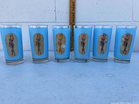 Six blue Key Club cocktail glasses lined up in a row, each featuring a vintage pin-up girl graphic inside a keyhole shape.