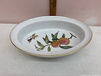 Oval white ceramic oven dish with painted peach fruit and flower motif, gold trim along rim, on white surface with ruler for scale