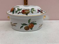 Side view of an oval Royal Worcester china casserole dish with fruit and wheat decoration and gold-accented handles and rim.