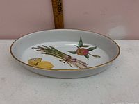 White ceramic oval serving dish with gold rim and fruit and asparagus painted decoration inside.