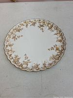 Top view of one white cake plate with gold floral decoration and scalloped edge.