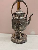 Front view of the Victorian ornate silver tip kettle with a hinged lid, curved spout, and detailed floral engravings, sitting on a round base stand.