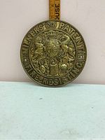 Close-up image of Victorian round brass safe plate embossed with Milners Patent Fire-Resisting, featuring a coat of arms and ornamental motifs, placed next to a ruler for scale.
