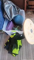 Weighted vest in black and neon green, blue exercise ball, purple yoga blocks, and small green dumbbells placed inside a plastic container.