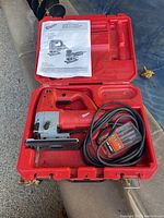 Full view of Milwaukee jig saw with power cord in open red plastic carrying case alongside instruction manual.