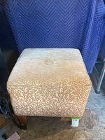 Front view of gold color Ethan Allen ottoman with patterned upholstery and wooden legs on stone floor.