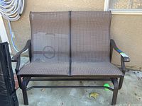Front view of a two-seat outdoor rocking bench with brown woven fabric seating and dark brown metal frame, placed on concrete near wall.