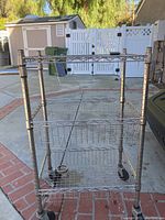 Metal rolling cart outdoors on brick and concrete surface showing front view with three wire mesh shelves and four caster wheels.