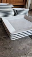 Close-up of stacks of white square plates emphasizing the size and shape, plates have slightly raised edges and appear to be ceramic or porcelain.
