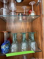 Wine glasses arranged on a glass shelf in a wooden cabinet. Bottom shelf displays six clear green glasses with embossed detailing and two colorful hand-blown style glasses.