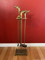 Full view of vintage brass fireplace tool set with three tools topped with horse head handles on brass stand against red wall