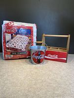 Photo showing all three Coca-Cola collectible items together: wooden condiment holder, metal tin bucket, and wrapped twin bed blanket.